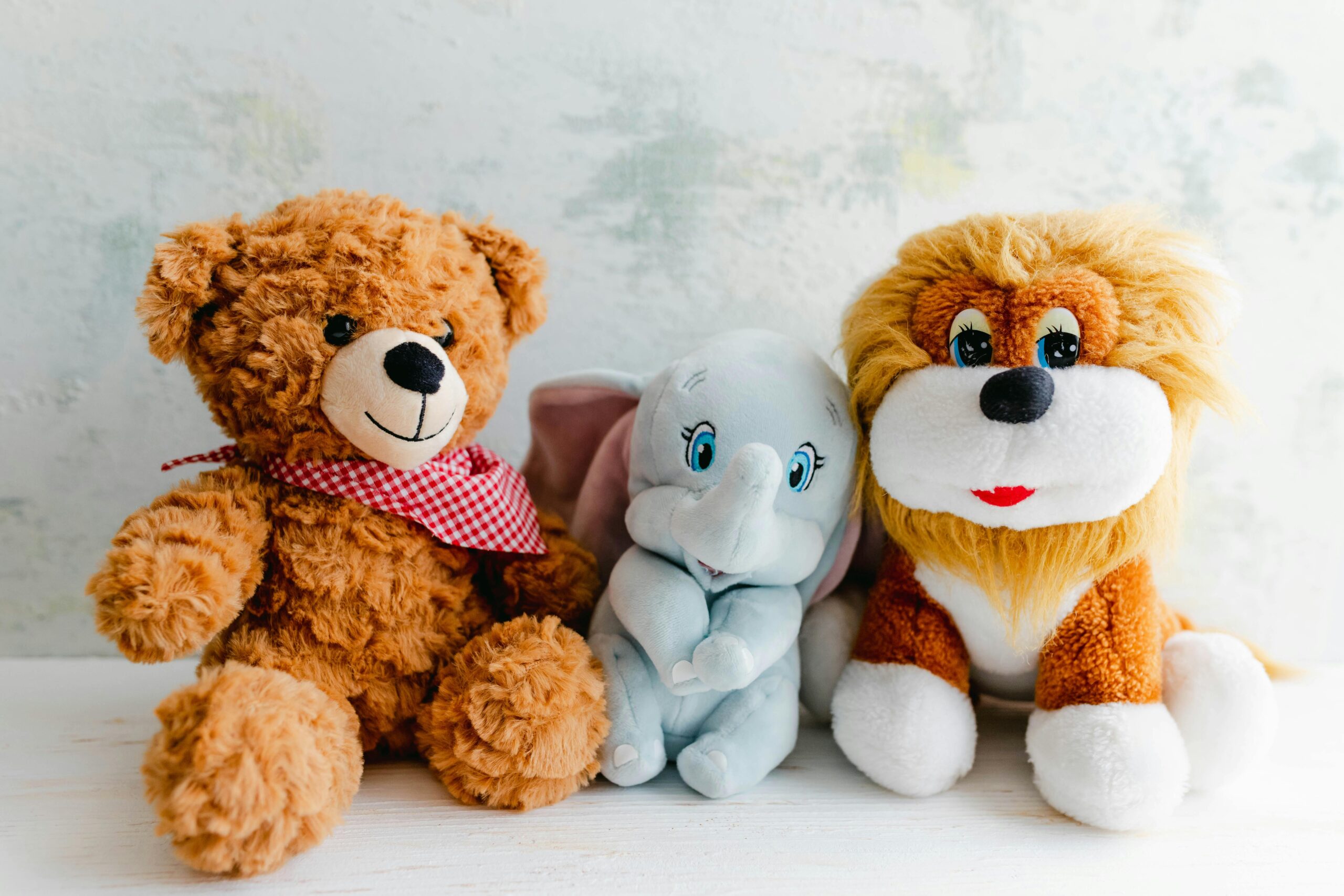 Zelienople Library Three plush toys—a brown teddy bear with a red checked scarf, a light blue elephant, and a lion—are sitting side by side on a white surface against a light background.